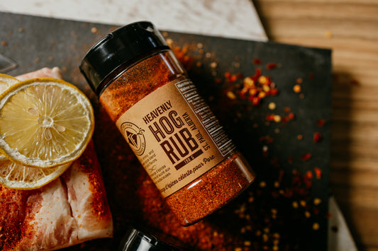 Bottle of 'Heavenly Hog Rub' spice mix with sliced lemon and pork on a wooden surface.