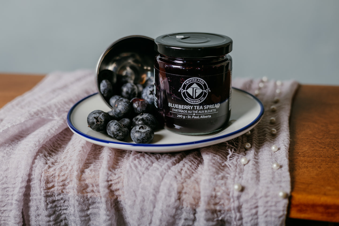 A jar of blueberry tea spread with its lid on a plate, the spread is served on a textured fabric with pearls scattered around.