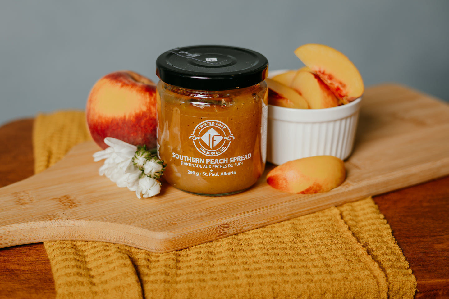 A jar of Southern Peach Jam with a label that reads 'Southern Peach Jams & Jellies, 290g', placed on a wooden board next to sliced peaches and a yellow cloth.