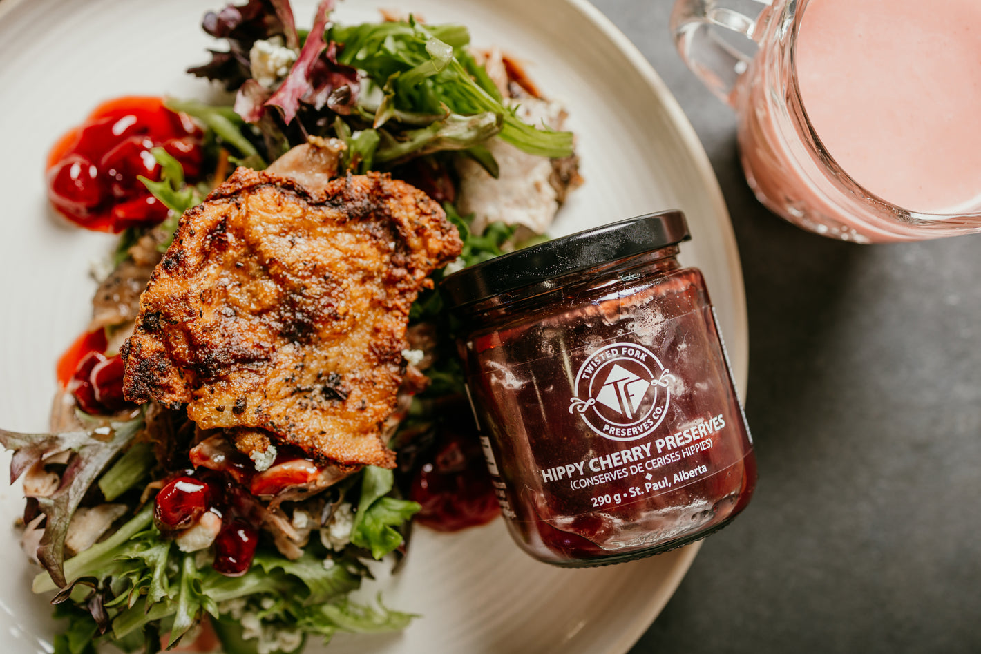 Plated dish with salad and fried item, accompanied by a jar of Hippy Cherry Pickles on a dark surface.
